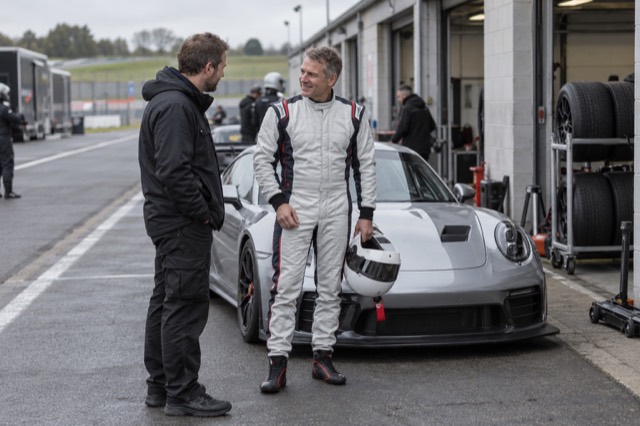 A club racing driver speaking with a mechanic beside a track car in the paddock
